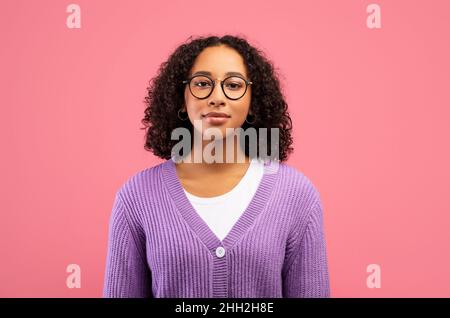 Positiva giovane donna nera che indossa occhiali e guarda la fotocamera, posando su sfondo rosa studio Foto Stock