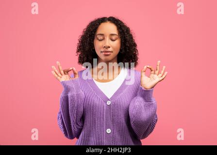 Gestione dello stress. Tranquilla giovane donna nera meditating, trovando l'equilibrio interno e l'armonia sullo sfondo rosa dello studio Foto Stock