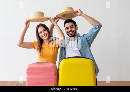 Vacanza in famiglia. Felice coppia turistica togliendo cappelli con gesto di saluto in piedi dietro bagagli valigia Foto Stock