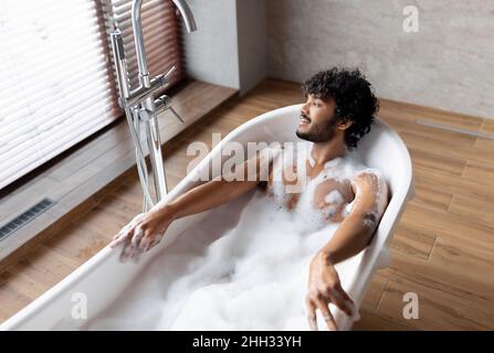 Home spa e relax nei fine settimana. Rilassato uomo indiano sdraiato in vasca da bagno con schiuma, godendo bagno bolla al mattino Foto Stock