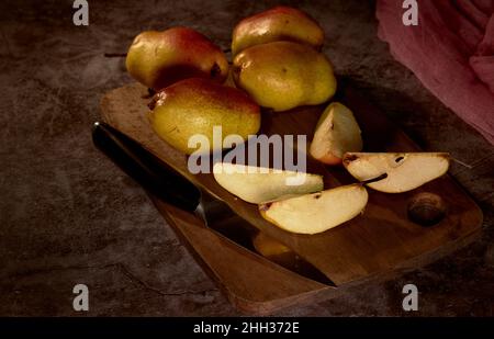 Tavola di pere. Le pere e un coltello sono su un tagliere e uno è tagliato in quattro parti e l'intera composizione su uno sfondo scuro Foto Stock