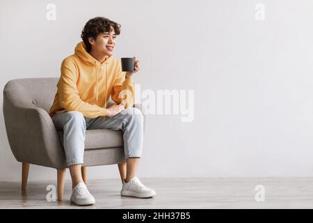 Bel giovane uomo asiatico che beve caffè, dopo una pausa, chillando in poltrona con bevanda calda contro la parete bianca studio Foto Stock