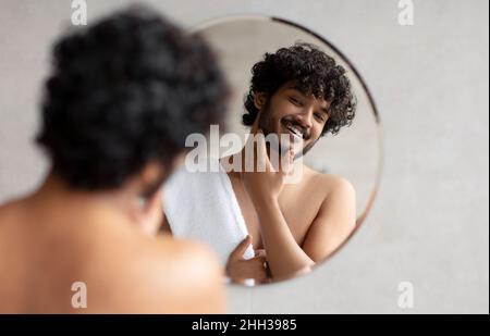 Giovane uomo indiano che guarda nello specchio in bagno e toccando la barba, bel ragazzo sorridente al suo riflesso Foto Stock