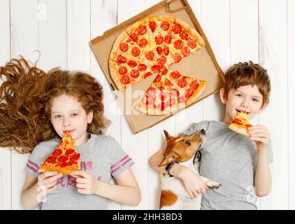 Bambini e cani che mangiano pizza al peperone. Bambini che tengono una fetta di pizza adagiata sul pavimento in legno. Festa di quarantena a casa. Foto Stock