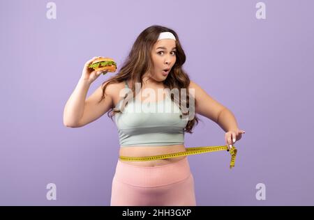 Sorpresa divertente affamato caucasico più taglia giovane donna in uniforme sportiva con hamburger misure vita con metro a nastro Foto Stock