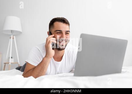 Felice ragazzo giovane attraente con barba si trova sul letto bianco chiamata dal telefono, guarda il portatile, lavoro o chat Foto Stock