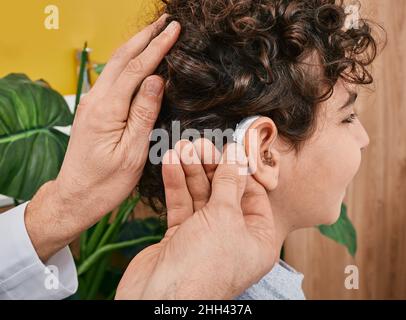 Montaggio di un apparecchio acustico sull'orecchio maschile del bambino presso la clinica uditiva, primo piano, vista laterale. Trattamento sordità, soluzioni uditive per bambini Foto Stock