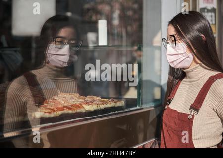 Giovane donna asiatica che indossa maschera facciale guardando una vetrina pizzeria. Foto Stock