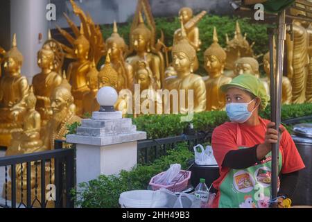 Un cuoco in una bancarella di cibo a Bangkok, Thailandia, guarda al suo fianco, un assortimento di statue di Buddha fatte in una fabbrica adiacente che fornisce un inusuale b/g Foto Stock
