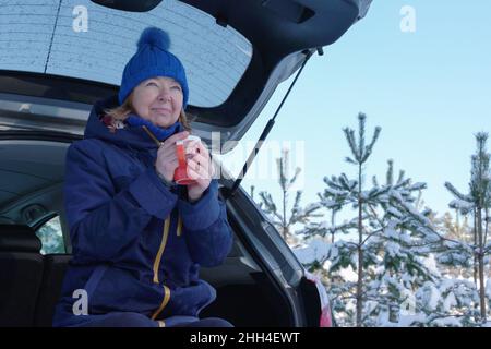 Sciatore maturo che ha il tè mentre si siede sul retro del suv dopo il sentiero della foresta di wintry. Attività all'aperto per aumentare l'umore durante la pandemia di coronavirus in inverno Foto Stock