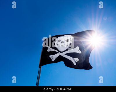 Bandiera nera dei pirati con un cranio bianco e ossa sul palo di legno bianco che ondola nel vento su sfondo cielo blu chiaro con raggi di sole luce. Foto Stock