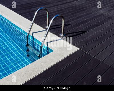 Cornice in pietra sulla piscina quadrata con acqua pulita, nessuno. Piscina a forma quadrata con piastrelle blu con maniglioni scala su legno scuro Foto Stock