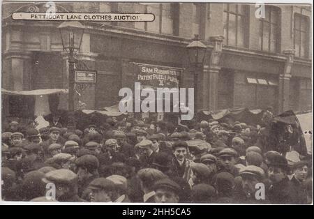 Petticoat Lane abbigliamento asta: Grande folla (per lo più uomini in cappe piatte) in strada, inizio 20th secolo. Un segno per il brevetto di Sam Rubinstein puzzle borses è sullo sfondo Foto Stock