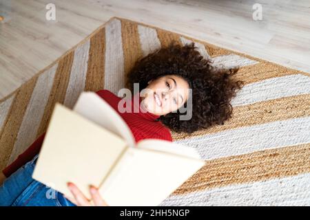 Giovane donna con libro sdraiato sul tappeto a casa Foto Stock