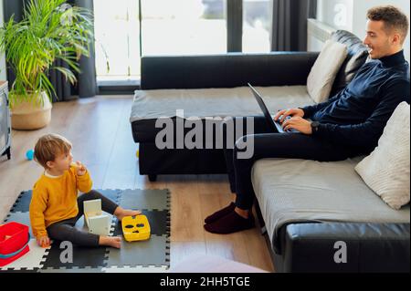 Bambino che guarda il padre che lavora sul computer portatile in salotto Foto Stock