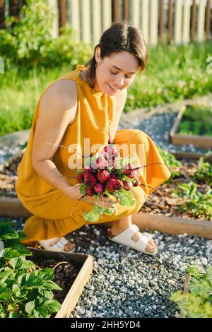 Donna sorridente che tiene un rafano appena raccolto in orto Foto Stock