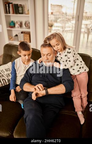 Ragazzo e ragazza seduti con nonno usando l'orologio intelligente seduto sul divano a casa Foto Stock