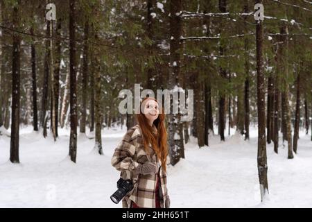 Donna rossa sorridente con macchina fotografica giorno sognando nella foresta invernale Foto Stock