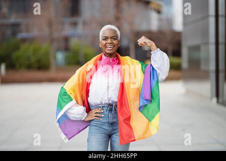 Giovane donna avvolta in bandiera arcobaleno muscolo flessibile con mano sull'anca Foto Stock