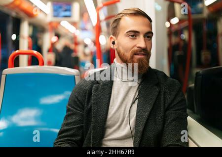 Giovane uomo che guarda fuori dalla finestra del tram e ascolta la musica attraverso le cuffie intrauricolari Foto Stock