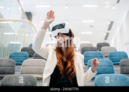 Donna da lavoro Redhead che indossa cuffie per realtà virtuale gesturando in ufficio Foto Stock