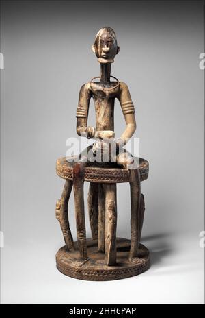 Figura: Uomo seduto su sgabello 18th–19th secolo (215+/-90 anni) popoli Dogon questa figura dominante posizionata al centro di uno sgabello circolare è definita dalla simmetria bilaterale enfatica e dalla chiarezza formale favorita dagli scultori Dogon. La composizione è notevole per l'elegante integrazione della figura e della struttura della sua sede che consiste di due dischi paralleli Uniti da un montante centrale e cinque supporti scolpiti. L’asse verticale del corpo superiore della figura è continuo con il montante centrale dello sgabello. Le sue ampie gambe allungate terminano in piedi saldamente piantati sulla superficie del suo b Foto Stock