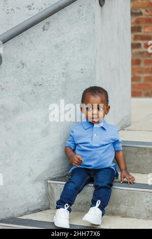 Un ragazzo carino afroamericano di un anno di età quasi prescolare con grandi occhi seduti sui gradini della città Foto Stock