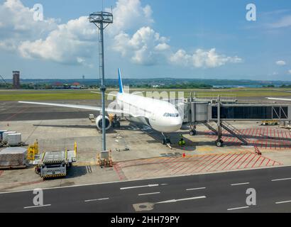 Giornata di sole all'aeroporto indonesiano. Un aeromobile con ponte jet collegato per l'imbarco e lo sbarco dei passeggeri. Oceano e giungla nel backgro Foto Stock