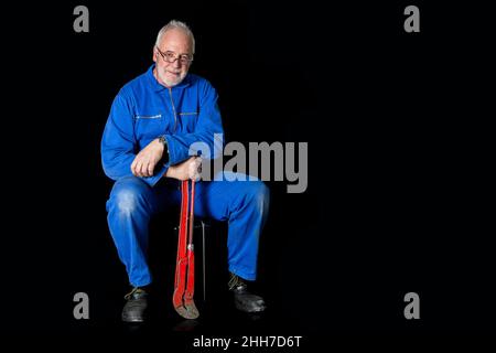 Un vecchio, esperto montatore in tuta blu si siede sulla sua custodia di montaggio su uno sfondo nero, tiene una grande chiave a tubo rossa nella sua mano. Foto Stock