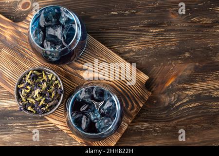Due bicchieri con tè al fiore di piselli farfalla e cubetti di ghiaccio su sfondo di legno, bevanda anchan blu alle erbe. Foto Stock