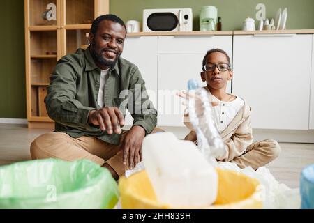 Ritratto a tutta lunghezza di felice padre afro-americano e figlio mettendo plastica in bidoni di riciclaggio a casa, spazio copia Foto Stock