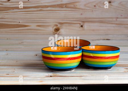 Fotografia di tre ciotole a strisce, colorate in rosso, blu, giallo, arancione e verde, su sfondo di legno; spazio di copia Foto Stock
