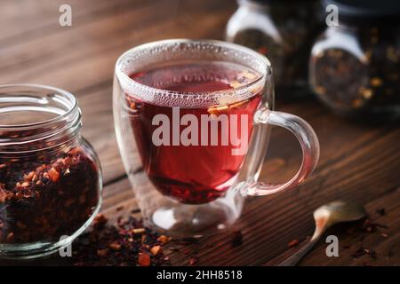 Bicchiere di tè caldo aromatico alla frutta rossa, vasetto di tè naturale alla frutta con petali di ibisco, fette di frutta e bacche. Vasetti di tè alla frutta aromatica sul dorso Foto Stock