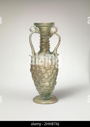 Bottiglia di vetro a forma di mazzo di uve 4th sec. d.C. romana incolore con colore verde chiaro; manici, piede e sentiero dello stesso colore.bordo arrotondato e leggermente addensato; bocca svasata; collo cilindrico alto; corpo ovoide allungato; anello largo applicato per il piede con bordo arrotondato rovesciato e fondo cavo; cicatrice pontil al centro del fondo; due maniglie attaccate alla parte superiore del corpo con aggetto schiacciato sporgente alla base, disegnate in una curva e tracciate sul collo (sopra la decorazione del sentiero) con un anello sopra, e terminanti sul bordo esterno del bordo. La cucitura verticale prominente dello stampo scorre lungo il corpo dall'alto verso il basso sotto h Foto Stock
