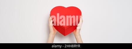 Mani che tengono grande scatola rossa a forma di cuore. Banner trendy per il giorno di San Valentino, il giorno Internazionale delle Donne o il giorno delle madri. Banner primo piano Foto Stock