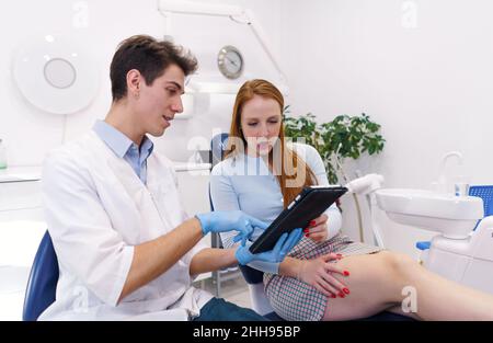 Giovane uomo in accappatoio bianco e guanti in lattice che mostrano i dati su tablet moderno a donna rossa mentre lavora in un moderno studio dentistico Foto Stock