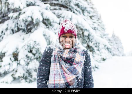 uomo afro-americano in cappello rosso ed elegante cappotto a plaid guardare la macchina fotografica con il sorriso bianco-neve al coperto nel parco Foto Stock