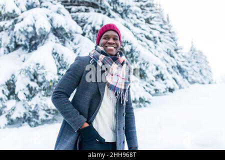 uomo afro-americano in cappello rosso ed elegante cappotto a plaid guardare la macchina fotografica con il sorriso bianco-neve al coperto nel parco Foto Stock