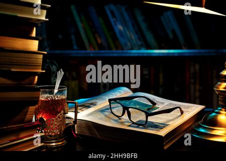 Libreria. Un libro aperto sotto una lampada da scrivania, con semplici occhiali neri. Un bicchiere di tè caldo. Sfondo del libro sugli scaffali. Foto Stock