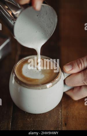 Versare il latte al vapore nel cappuccino in tazza bianca contro il tavolo di legno scuro. Moody Coffee Shop Lifestyle. Piatto caffè con spazio copia. Cortado. Piatto Foto Stock