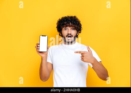 Shocked sorpreso indiano ragazzo in t-shirt bianca, tiene smartphone con mock-up bianco schermo vuoto e punta il dito su di esso, si leva su isolato sfondo arancione, scioccato guarda la fotocamera Foto Stock