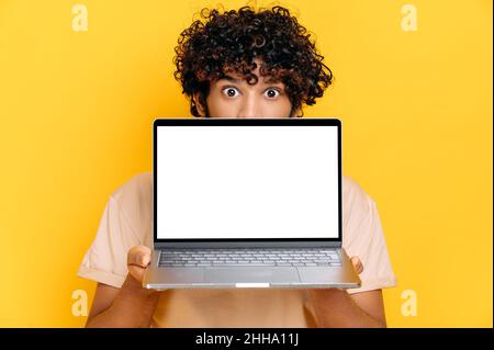 Stupito il giovane indiano eccitato che si alza da dietro il laptop, guarda sorpreso la fotocamera, si erge su sfondo arancione isolato, tiene un notebook aperto con schermo bianco vuoto, spazio copia, mock-up Foto Stock