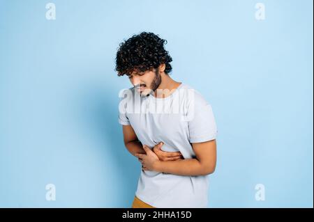 Mal di stomaco. Il ragazzo indiano con in abiti casual che appiccano e tengono il suo ventre, soffre di dolore all'addome o allo stomaco, ha avuto avvelenato, ha bisogno di trattamento, si leva su sfondo blu isolato Foto Stock