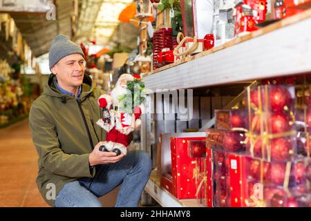 Prepararsi per il nuovo anno - l'uomo felice sceglie una figurina di babbo natale nel magazzino del negozio Foto Stock