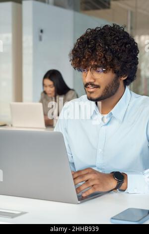 Il giovane e vivace uomo indiano in occhiali lavora su un computer portatile seduto alla scrivania Foto Stock