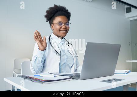Sorridente giovane infermiera femminile in divisa medica seduta alla scrivania ha videocall sul laptop Foto Stock