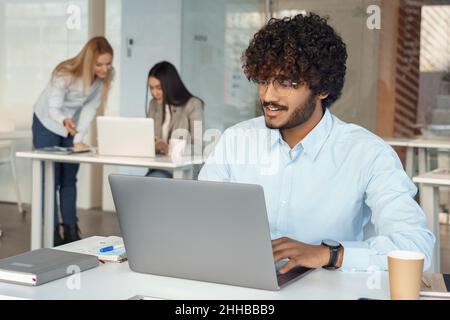 Un uomo indiano impegnato lavora su un laptop alla scrivania mentre un collega si spavesta in background Foto Stock