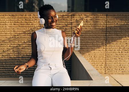 Donna nera sorridente mentre ama ascoltare la musica con un telefono cellulare e le cuffie all'aperto. Foto Stock