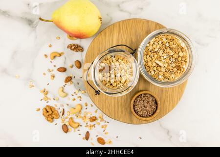 vista dall'alto di due vasetti di vetro con muesli o granola su un asse di legno e un tavolo di marmo con noci e uvetta sparse. corretta alimentazione Foto Stock