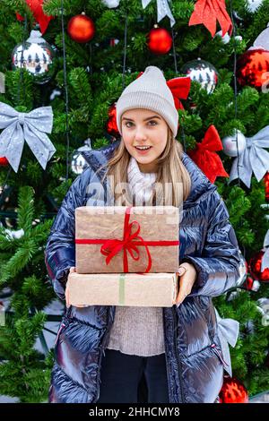 Ragazza bionda carina con regali di Natale sullo sfondo di un albero di Natale decorato. Guardando la fotocamera. Scatto verticale Foto Stock